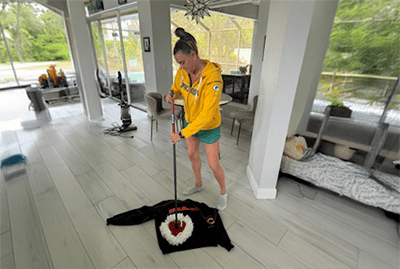A woman in a yellow hoodie uses a mop on a black shirt with a white collar in a bright, modern living space. The scene illustrates a cleaning activity.